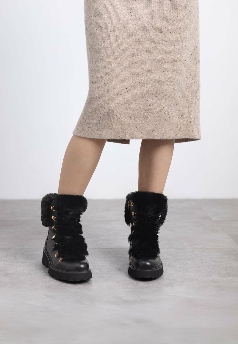 Botas de tobillo forradas de piel sintética negra con suela de goma gruesa, ojales de metal y un diseño de cordones. La parte superior presenta un acabado suave y texturizado.