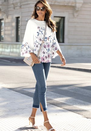 Witte bloemenblouse met belmouwen, blauwe jeans met zijslits en nude strappy hakken; model loopt op een stadsstraat.
