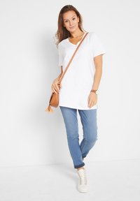 T-shirt bianca con scollo a V, vestibilità morbida, abbinata a jeans blu. Borsa a tracolla marrone piccola, sneakers bianche e un orologio con cinturino marrone.