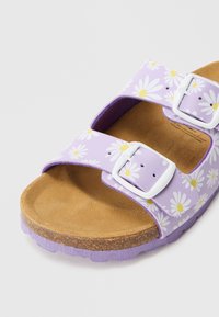 Fialová sandála s potlačou bielych margarét, vybavená dvoma nastaviteľnými remienkami s bielymi prackami a hnedou semišovou stielkou. Textúrovaná fialová podrážka.