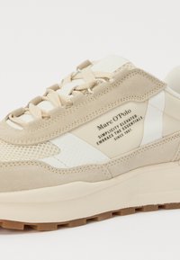 Beige sportcipő textúrázott velúrból és hálós anyagból, fehér kiegészítőkkel és vastag gumitalppal. A márkajelzés a cipő oldalán látható.