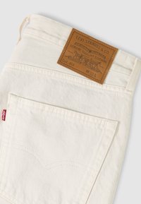 Λευκή πίσω τσέπη τζιν Levi’s με καφέ δερμάτινο λογότυπο της μάρκας και κόκκινη υφασμάτινη ετικέτα Levi’s στη ραφή της πλευράς.