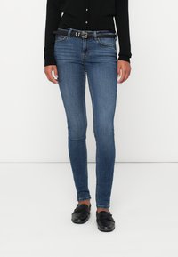 Moteris, vilkinti siaurus mėlynus džinsus su juodu diržu, juodą megztuką su mygtukais ir juodais loaferiais, stovi prieš baltą foną.