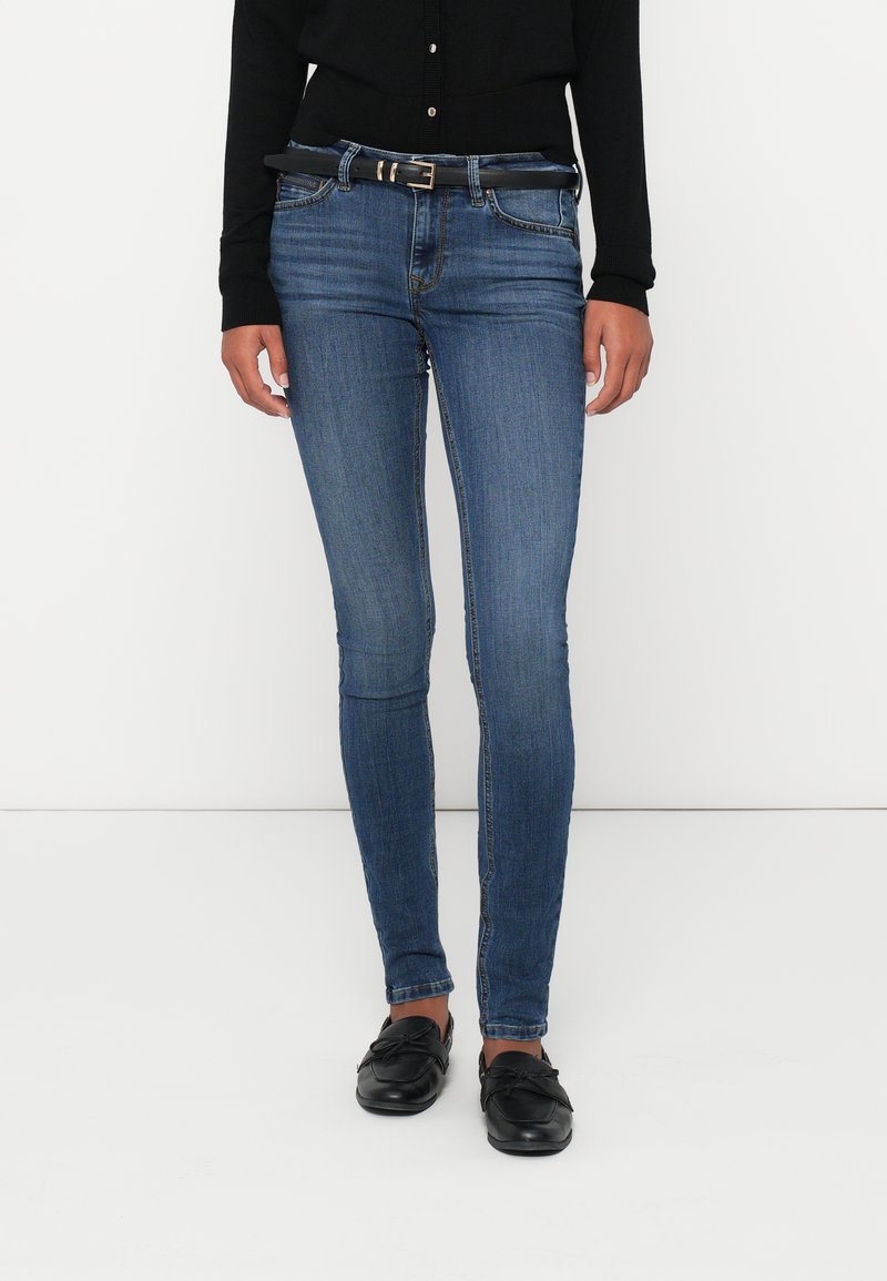 Moteris, vilkinti siaurus mėlynus džinsus su juodu diržu, juodą megztuką su mygtukais ir juodais loaferiais, stovi prieš baltą foną.