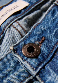 Tessuto di denim blu con cuciture visibili, caratterizzato da un bottone in metallo con marchio inciso e un'etichetta di tessuto applicata.