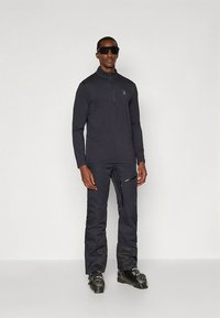 Svart, langermet zip-up genser og matchende bukser, laget av stretchstoff; har en glidelåslomme og glatt tekstur; brukt med skistøvler.