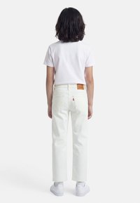 Παιδί με σκούρα μαλλιά μέχρι τους ώμους, που φοράει άσπρο κοντομάνικο μπλουζάκι, άσπρα τζιν και άσπρα αθλητικά παπούτσια, στέκεται με την πλάτη.