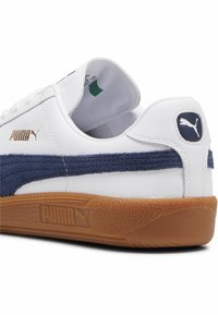 Weiße Ledersneaker mit einem Marineblauen Wildlederstreifen, einer Gummisohle in Kaugummi-Optik und goldfarbenen Logo-Akzenten an der Seite und Ferse.