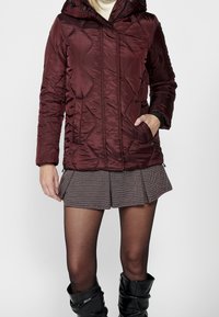 Doudoune bordeaux avec capuche, présentant un design matelassé, des poches zippées et une texture lisse. Associée à une jupe à carreaux et des bottes montantes jusqu'aux genoux.