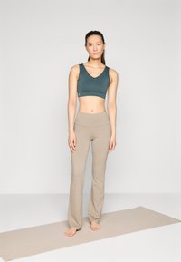Sostén deportivo en verde oscuro con escote en V, combinado con pantalones de yoga de talle alto en beige. Textura suave, diseño minimalista, de pie sobre una esterilla de yoga.