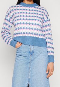 Mujer vistiendo un suéter de punto con un patrón en azul pastel, rosa y blanco, y una falda de mezclilla azul clara con una abertura frontal.