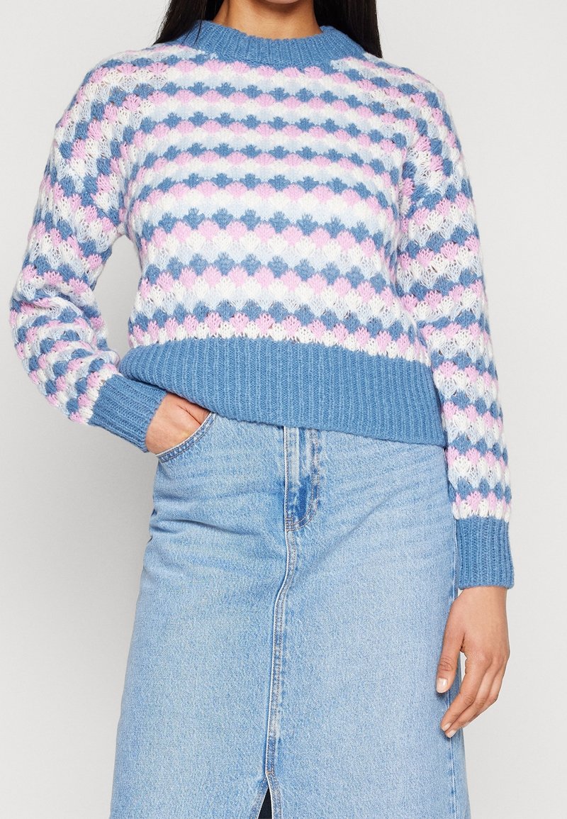 Mujer vistiendo un suéter de punto con un patrón en azul pastel, rosa y blanco, y una falda de mezclilla azul clara con una abertura frontal.