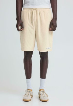 Mężczyzna ubrany w beżowe szorty z frotté materiału z troczkami, białe skarpety za kostkę i beżowe trampki, stojący na jednolitym tle.
