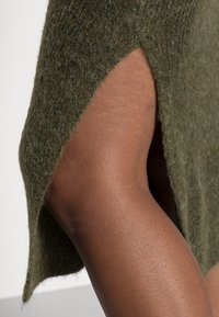 Grön stickad tröja med avslappnad passform, sidospårdetalj och mjuk textur som blottar hudtonen vid fållen.