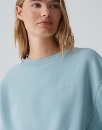 Sweatshirt azzurro chiaro realizzato in un tessuto morbido, con scollatura a coste e un piccolo ricamo a forma di cuore sul petto. Texture liscia.