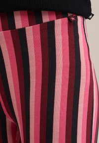 Csíkos leggings rózsaszín, piros és fekete árnyalataiban. Pamuthoz hasonló anyag, bordázott textúrával, kontrasztos derékkal és kis kiegészítő címkékkel.