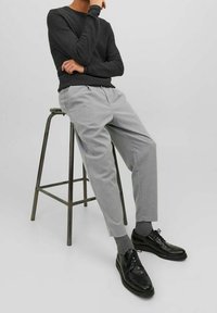 Grå strukturerade byxor och en svart långärmad skjorta, matchat med svarta läderskor och grå strumpor, sittande på en metallpall.