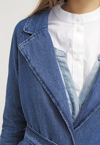 Jeansová bunda v tmavě modré barvě s velkým límcem a zúženým pasem. Přes světlou modrou košili s límečkem, která ukazuje kontrastní textury.