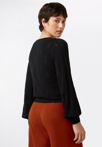 Schwarzer Strickpullover mit langen Ärmeln, der ein strukturiertes Pfeilmuster aufweist. Kombiniert mit strukturierten, rostfarbigen Hosen.