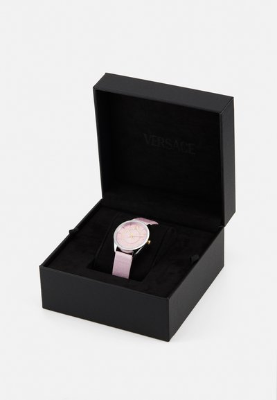 Versace watch in a black box. Features a silver case, pink dial, gold accents, and a pink fabric strap. Displayed on a black cushion.