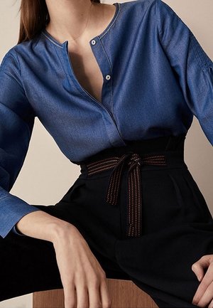 Personne portant une chemise bleue boutonnée rentrée dans un pantalon noir taille haute avec une ceinture en tissu nouée, assise avec une main reposant sur une surface en bois.