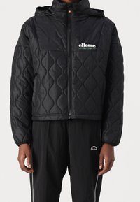 Czarna pikowana kurtka z kapturem, z zapięciem na zamek i logo "ellesse Ski Club" w zielonym kolorze. Miękka, gładka faktura z krojem cropped.
