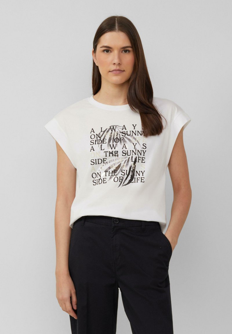 Witte katoenen t-shirt met opgerolde korte mouwen, met zwarte en gouden tekst en een grafisch ontwerp in het midden. Gecombineerd met donkere broek.