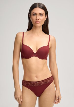 Femme portant un soutien-gorge rouge foncé assorti avec des bretelles fines et une culotte bordée de dentelle, debout devant un fond clair uni.