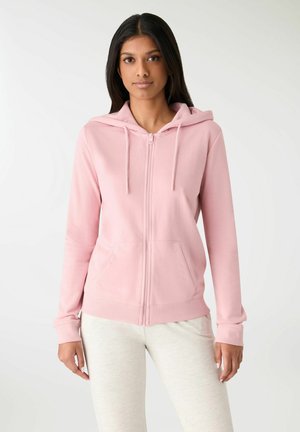 Donna che indossa una felpa con cappuccio rosa chiaro con zip, tasche frontali e coulisse, abbinata a pantaloni chiari, in piedi davanti a uno sfondo semplice.