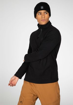 Zwarte fleece pullover met hoge kraag en rits, naast bruine cargobroek. Kenmerkt zich door tonale logo-details en lange mouwen.