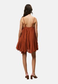 Rostfarbendes, ärmelloses Kleid mit einem schwingenden Rock und einem tiefen Rückenausschnitt. Hergestellt aus leichtem Stoff, mit dünnen Trägern und einem fließenden Design.