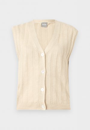 Γυναίκα με μπεζ αμάνικο γιλέκο με κουμπιά πάνω από ριγέ ζιβάγκο, φαρδιά πλισέ κρεμ παντελόνια και μυτερά τακούνια, στέκεται με τα χέρια πίσω.