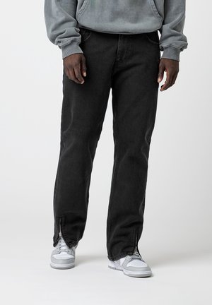 Personne portant un jean noir avec des fermetures éclair aux chevilles, des baskets blanches et grises, et un sweat gris, debout devant un fond uni.
