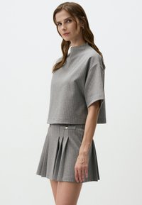 Graues Crop-Top mit hohem Hals und kurzen Ärmeln, kombiniert mit einem plissierten Rock. Beide Teile haben dünne vertikale Streifen und metallische Akzente.