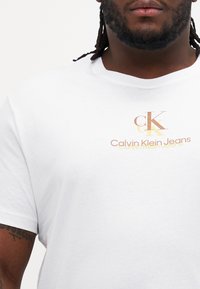 Baltas medvilninis marškinėliai su trumpomis rankovėmis, puošti auksiniu ir rudu spaudiniu „Calvin Klein Jeans“ ir „ck“ centre.