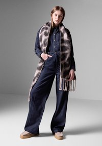 Tummansininen denim-haalaripuku, jossa on istuva yläosa ja leveät lahkeet. Pukeutunut beigen ja harmaan fringed-huivin kanssa, jossa on pilkullinen kuvio.