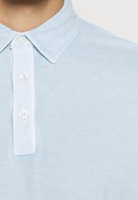 Vaaleansininen polo-paita, jossa on napitettava kaulus. Paita on pehmeätekstuurinen ja siinä on neljä nappia kauluksessa, mikä luo yksinkertaisen ja klassisen muotoilun.