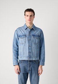 Light blue denim jacket with button closure, two chest pockets, and long sleeves. Worn over a light beige collared shirt.