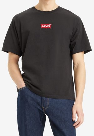 Personne portant un t-shirt noir Levi's avec un logo rouge sur la poitrine et un jean bleu, main dans la poche, sur fond blanc.