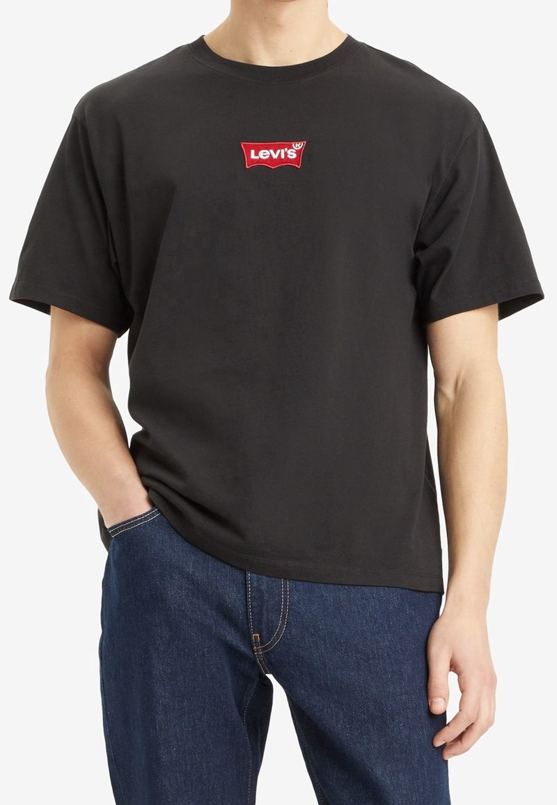 Personne portant un t-shirt noir Levi's avec un logo rouge sur la poitrine et un jean bleu, main dans la poche, sur fond blanc.