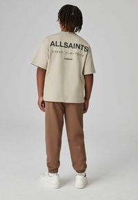 Beige lyhythihainen t-paita, jossa on musta teksti "ALLSAINTS" ja japanilaiset merkit selässä, yhdistettynä ruskeisiin collegehousuihin ja valkoisiin lenkkareihin.