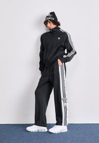 Tuta sportivo nero con strisce bianche e bottoni a scatto sui pantaloni. Completato con un berretto nero e scarpe sportive bianche.