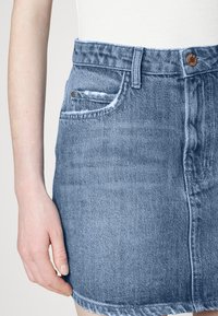 Saia de ganga em azul claro, com uma bainha desfiada, design clássico de cinco bolsos e fecho em botão metálico na cintura.