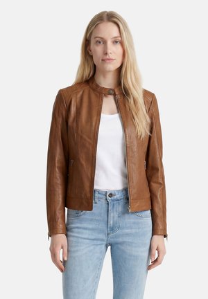 Frau mit langen blonden Haaren, die eine braune Lederjacke über einem weißen Hemd und hellblauen Jeans trägt, stehend vor einem einfarbigen Hintergrund.