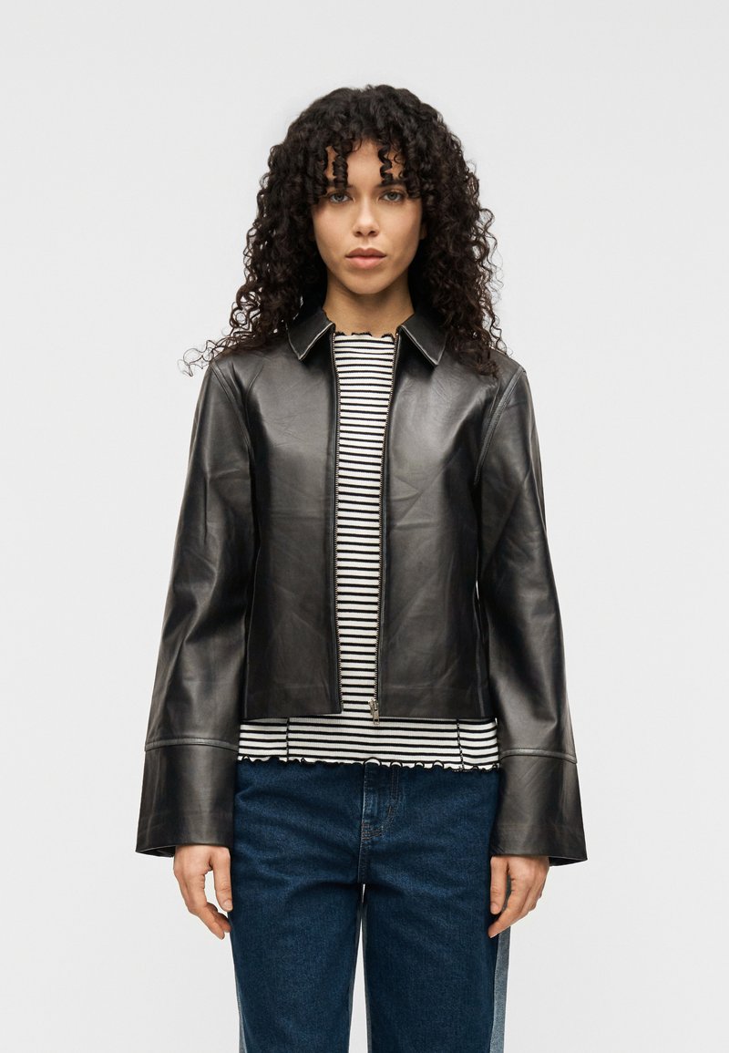 Μαύρο δερμάτινο μπουφάν με γιακά και κλείσιμο με φερμουάρ. Διαθέτει μανίκια σε σχήμα καμπάνας και ραφές με λεπτομέρειες. Φορεμένο πάνω από ριγέ τοπ.