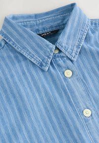 Chemise en denim bleu clair avec un col classique, une texture verticale et trois boutons blancs. Étiquette de la marque : NEXT.