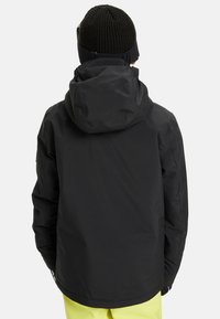 Osoba ubrana w czarną kurtkę z kapturem, czapkę z dzianiny w kolorze czarnym i żółte spodnie, stojąca tyłem na jednolitym jasnoszarym tle.