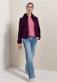 Veste en faux daim violet avec un col en fourrure, portée sur un pull rose et un jean flare bleu clair. La mannequin se tient devant un fond neutre.