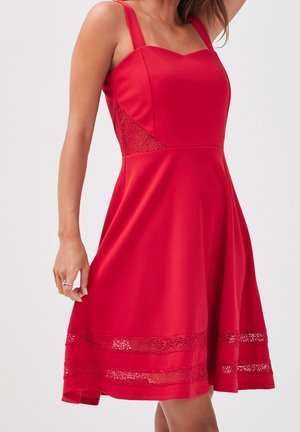 Femme portant une robe rouge sans manches avec une insertion en dentelle à la taille et deux bandes de dentelle près de l'ourlet, debout devant un fond uni.