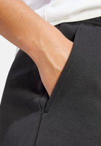 Pantalones de chándal negros con un bolsillo lateral, hechos de un tejido suave y texturizado. La toma en primer plano muestra una mano descansando en el bolsillo, enfatizando el diseño casual.